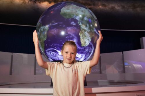 Young female Life Science Centre visitor stood in front of The Sphere, with arms outstretched as if they are holding the Earth.