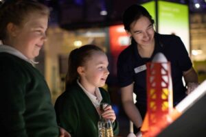 Two school students enjoying a rocket building activity with the support of an Explainer inside Life Science Centre.
