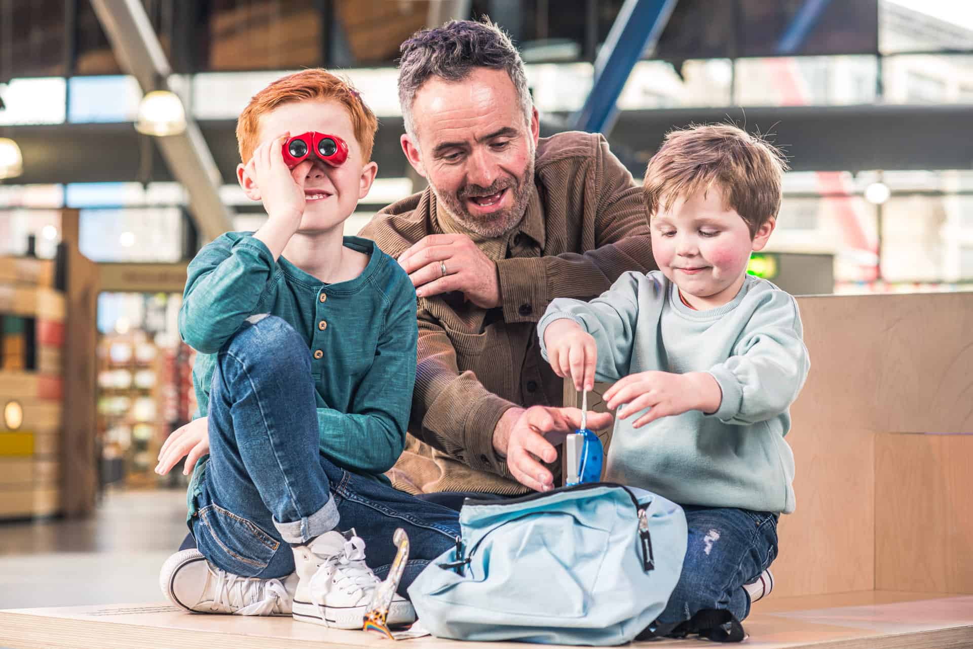 Father with two young sons looking through a Discovery Backpack inside Life Science Centre. One boy is looking through bright red binoculars.