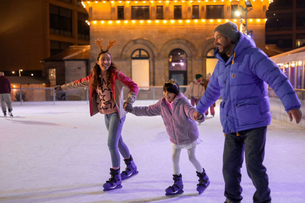 Family of three wrapped up for winter holding hands whilst skatings on Life Science Centre in Newcastle's outdoor ice rink.