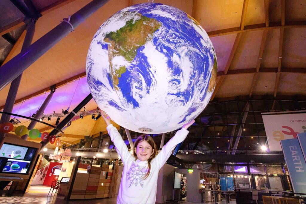 Young girl with outstretched arms as if she is holding Gaia, Life Science Centre's replica of Planet Earth.