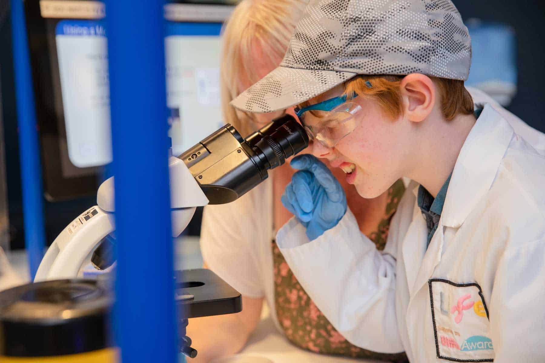 Young boy wearing a camouflage cap and looking through a microscope assisted by him mum in Life Science Centre's Experiment Zone.
