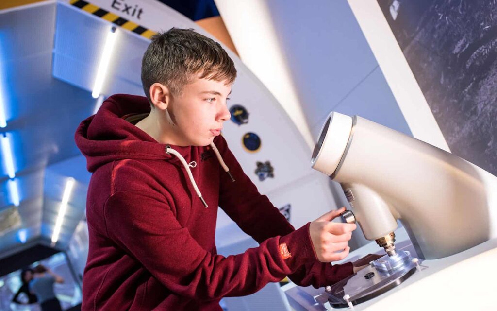 Male teenage visitor wearing a red hoody looking through a microscope in Life's Space Zone.