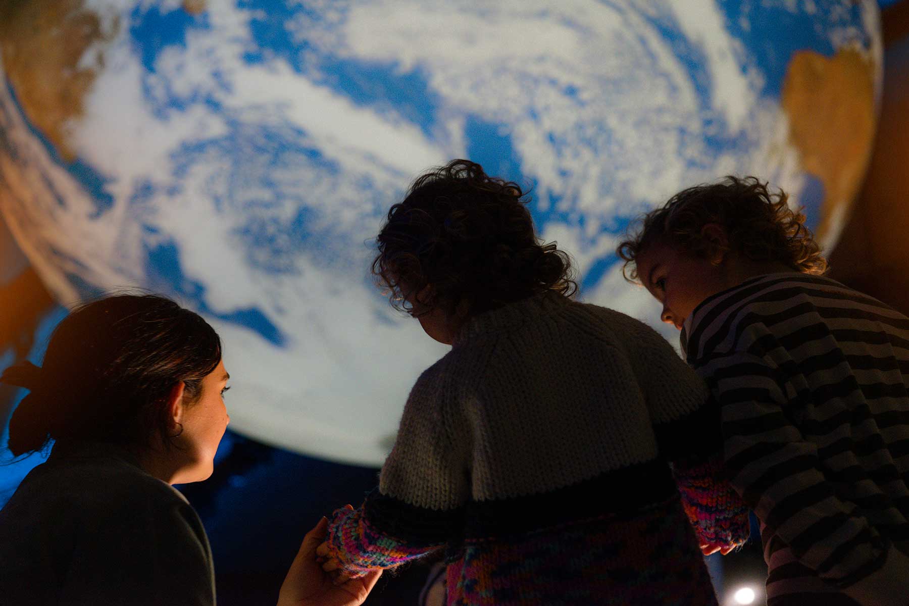 Mother with two children in dimly lit science centre looking up at Gaia, a large replica of Planet Earth