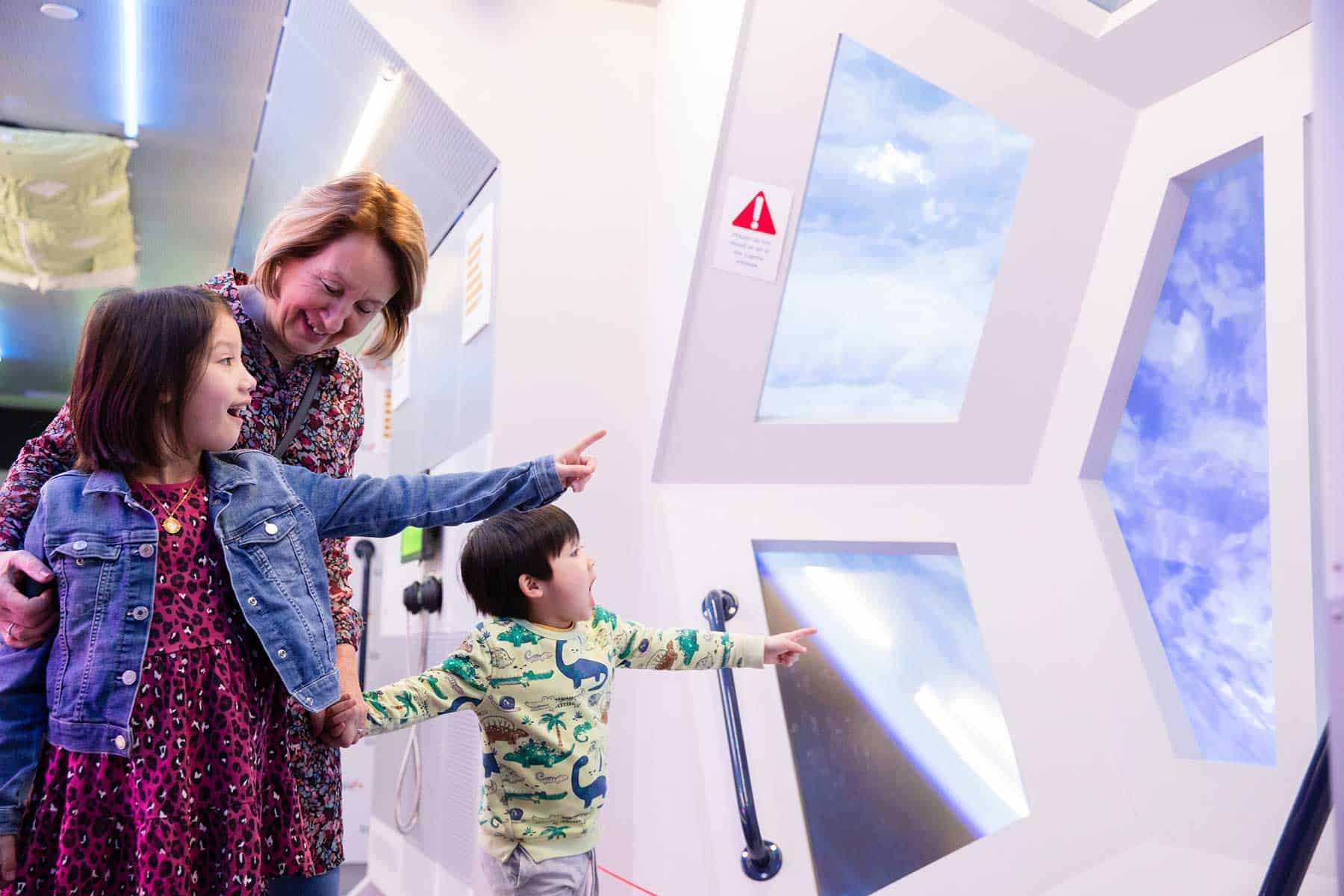 Grandmother with two children looking and pointing through ISS replica window in Life Science Centre's Space Zone.
