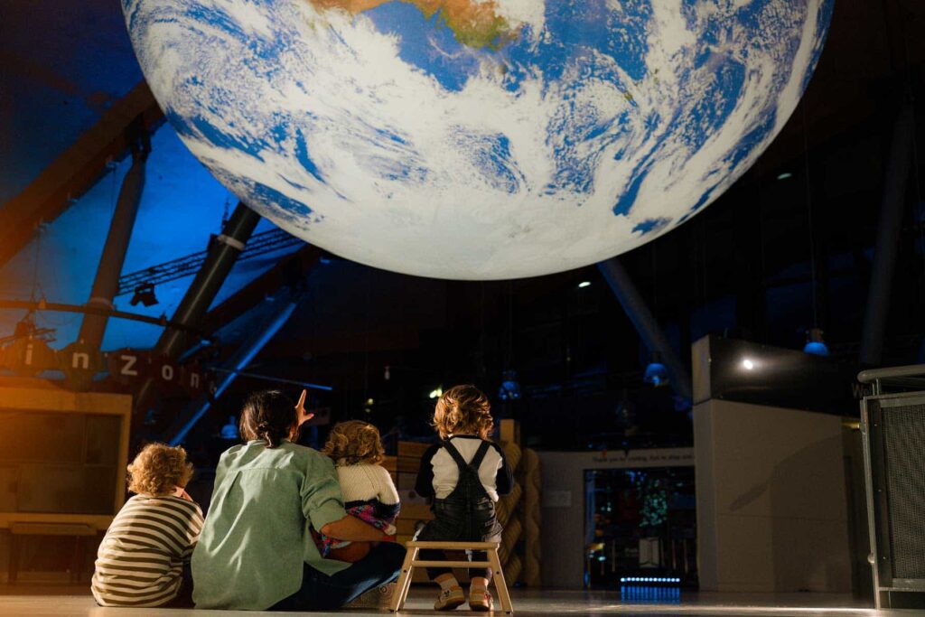 Family sat down in dimly lit science centre, looking up and pointing at Gaia, a glowing replica of Earth.