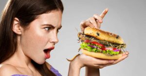 Lady with brown hair looking surprised with a burger containing a cricket in front of her