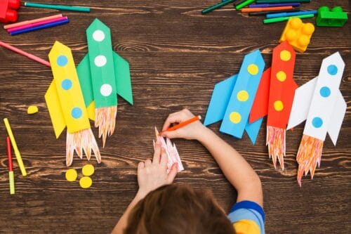Preschool Child In Creativity In The Home. Happy Kid Makes Rockets From Paper. Children's Creativity. Creative Children Play With Craft. Tools And Materials For Children's Art Creativity On Table.