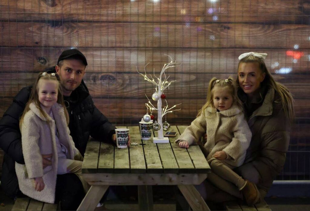 Family enjoying hot chocolates and smiling at camera on outdoor bench at Newcastle's ice rink.