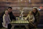Family enjoying hot chocolates and smiling at camera on outdoor bench at Newcastle's ice rink.