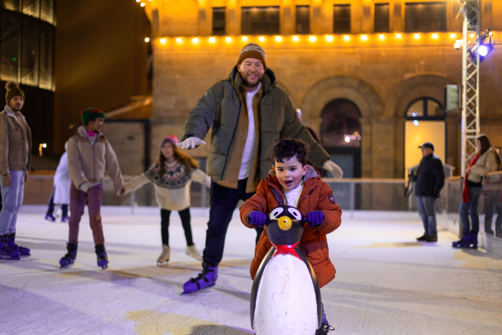 Little boy using skating aid on Life Science Centre's outdoor ice rink accompanied by father.