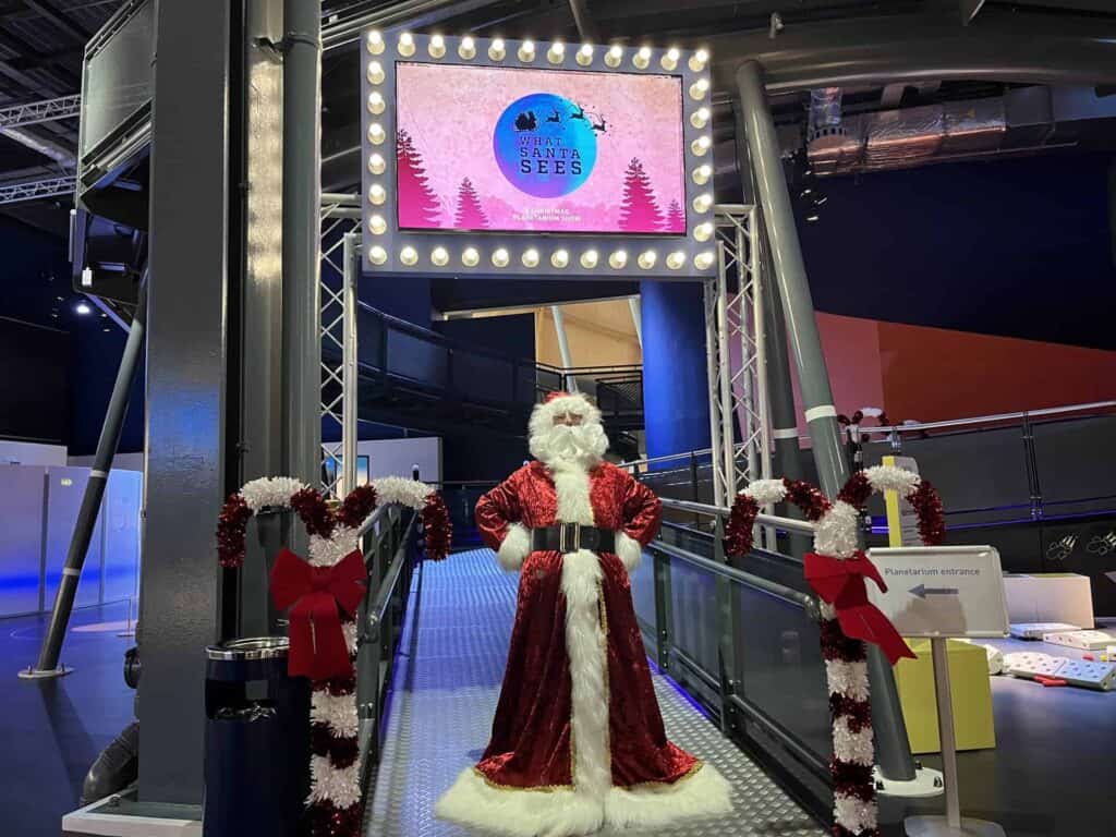 Santa in front of Life Science Centre's planetarium with 'What Santa Sees' screen in background.