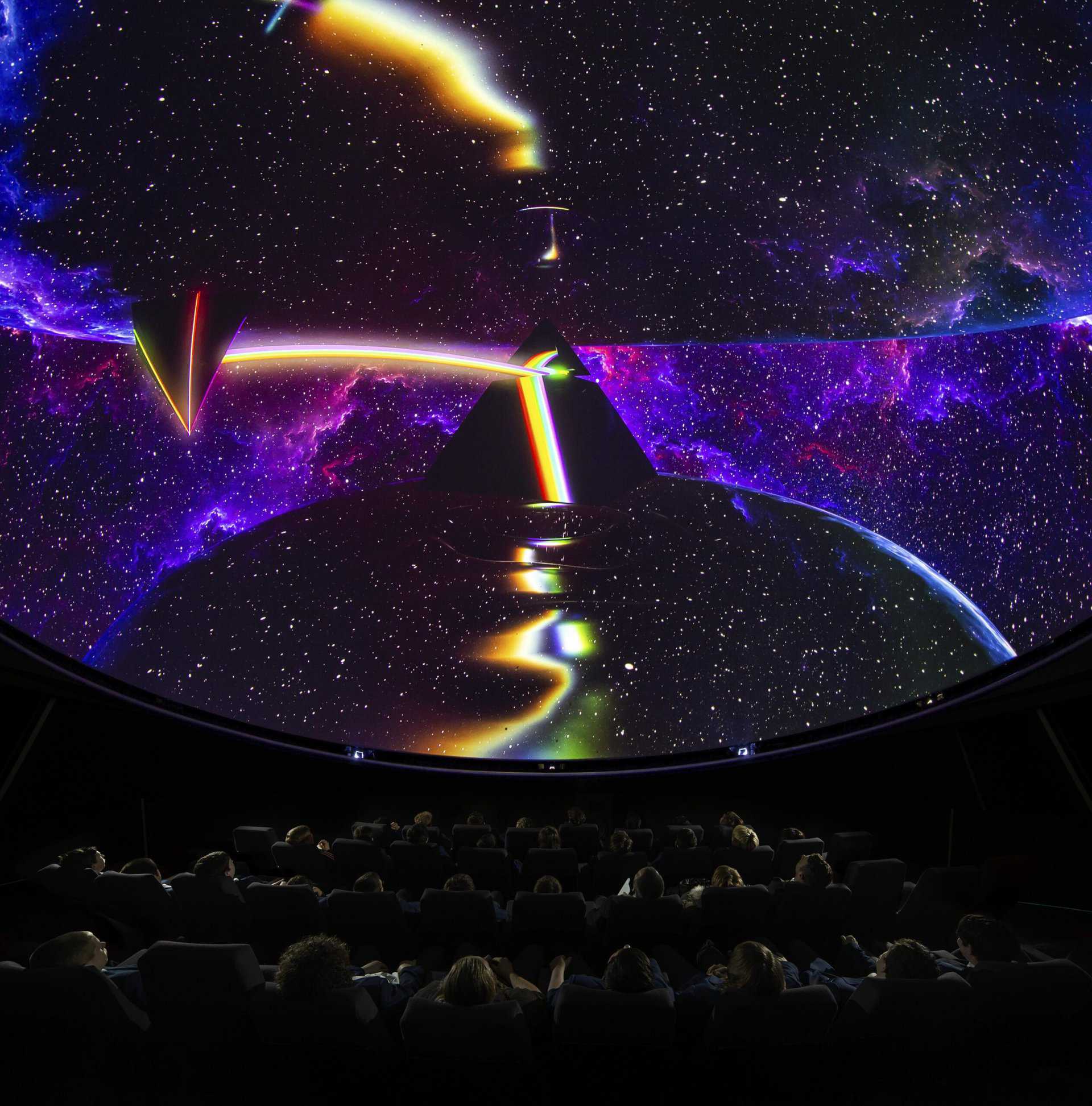 A group of adults, seen from behind, view an impressive rainbow- and space-themed visual on the planetarium dome screen.