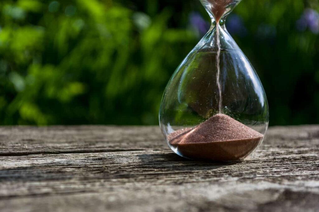 Hourglass with pouring sand against a background of blurred greenery. The bottom of a glass flask with toit on an old wooden table.