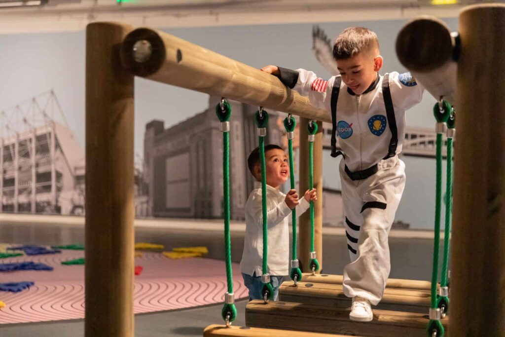 Young visitors in Life Science Centre's Play Zone. Older boy is crossing wooden bridge in space outfit