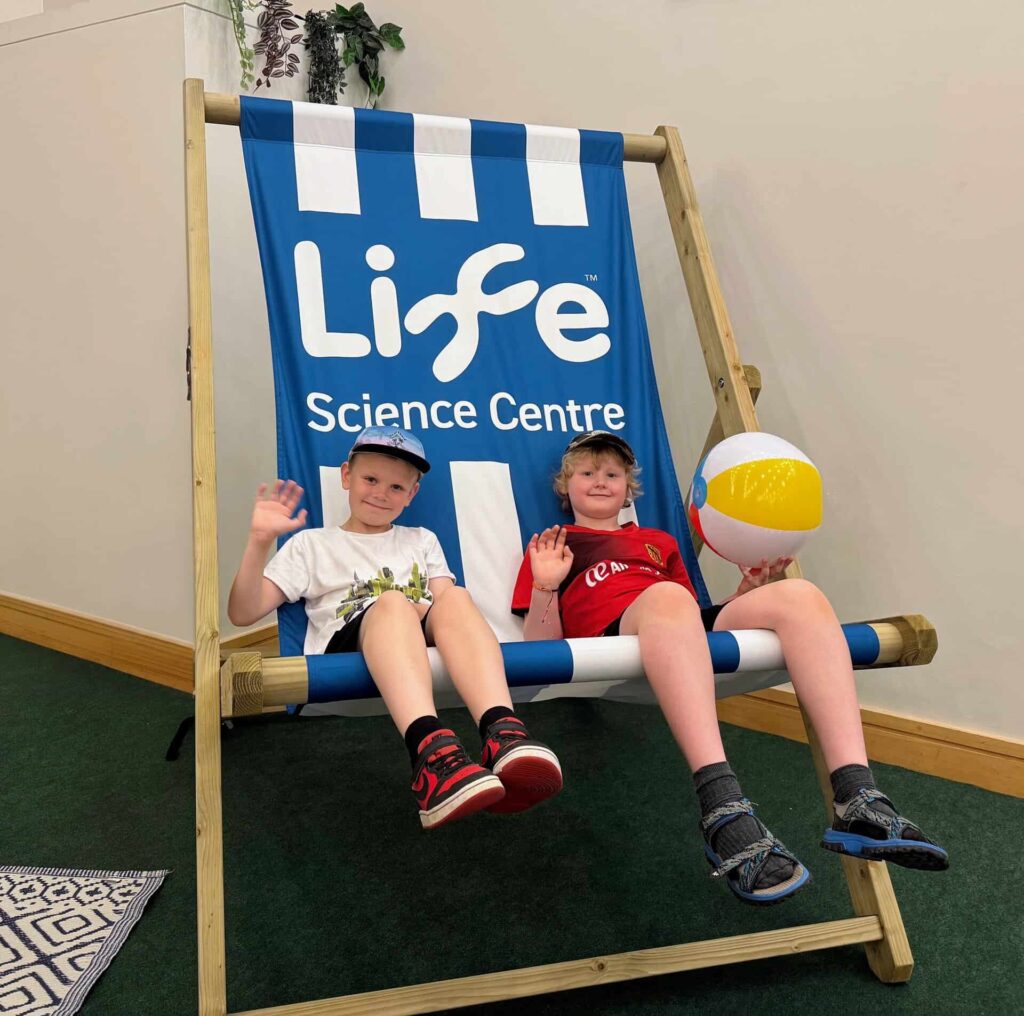 Two young boys sat on giant Life branded deckchair in science centre.