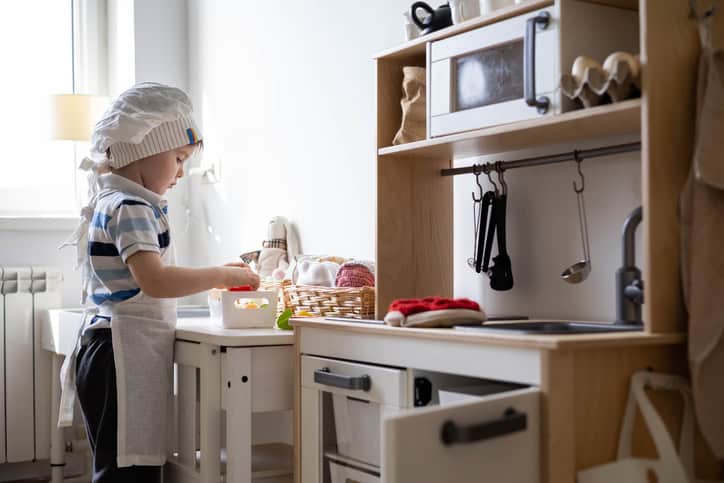 Cute little baby boy in chef hat and apron playing at childish kitchen cooking food back view. Male kid taking cups for preparing table tea drinking at cuisine playroom. Early development education