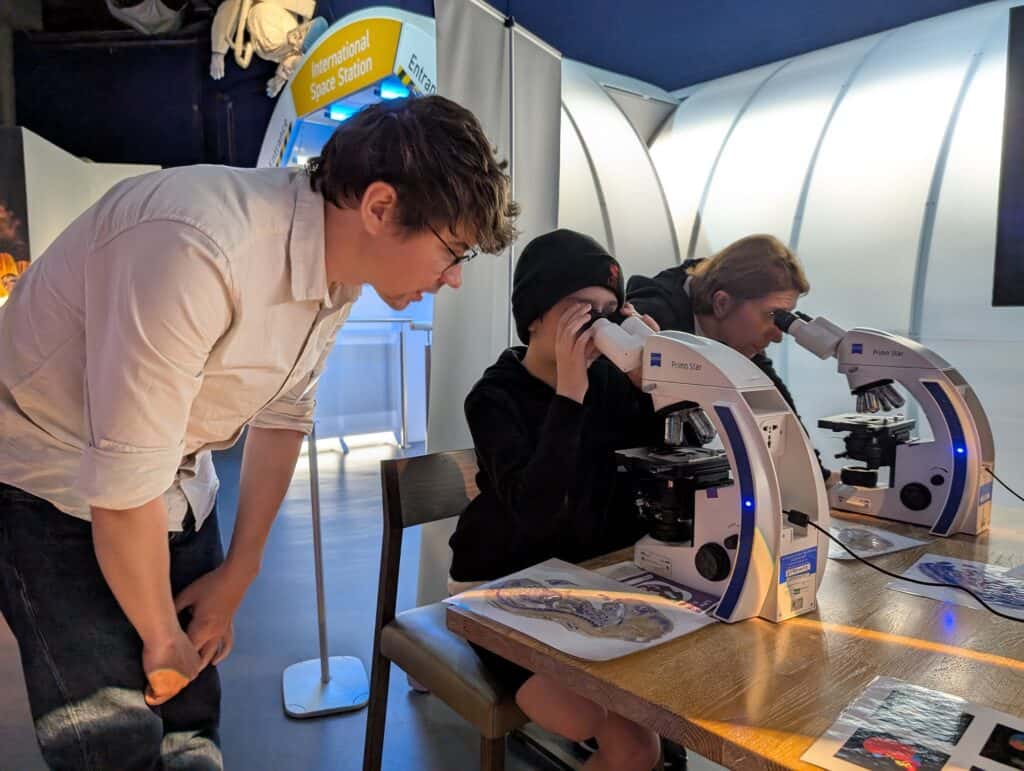 Little Boy Looking Through Microscope In Life Science Centre Space Zone, Assisted By Local Scientist.