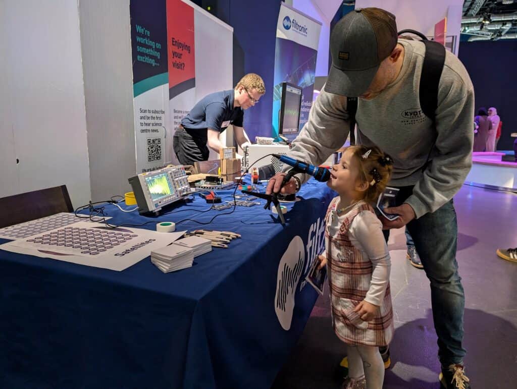 Little girl talking in mic with assistance of dad wearing a cap in science centre.
