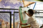 Little boy in white t shirt drawing planet Earth on an easel, looking out on Gaia, a replica of Earth inside Life Science Centre