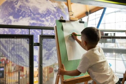 Little boy in white t shirt drawing planet Earth on an easel, looking out on Gaia, a replica of Earth inside Life Science Centre