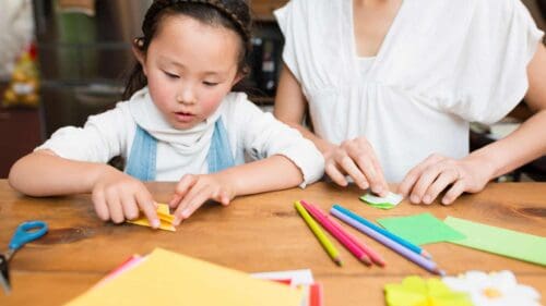young girl sitting at table with parent, playing with colourful paper.