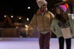 Two girls linking arms whilst skating on outdoor ice rink.