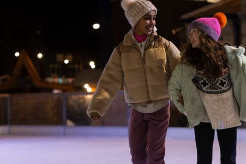 Two girls linking arms whilst skating on outdoor ice rink.