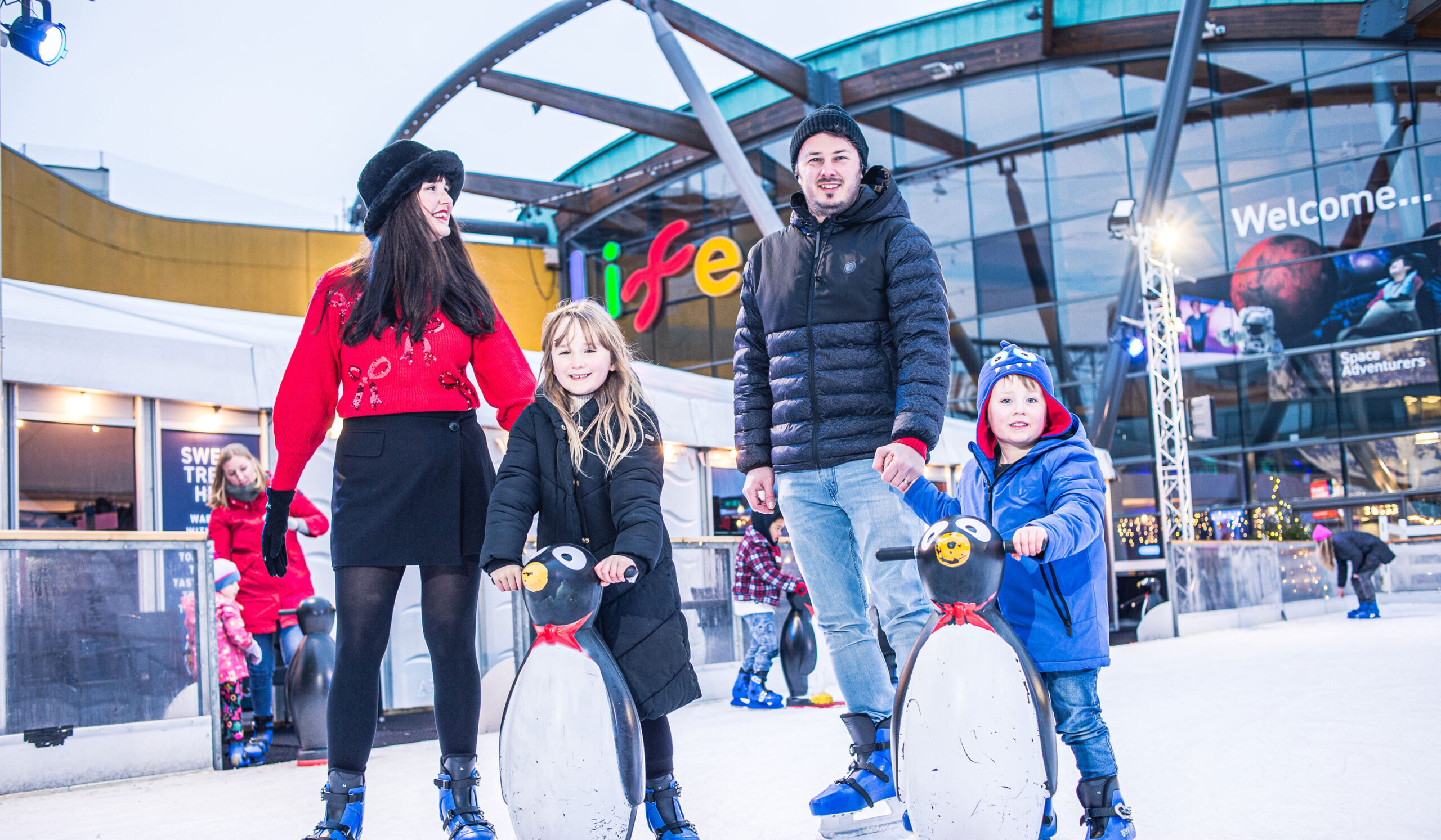 Family of four enjoying a skate on Life Sceince Centre's outdoor ice rink with children using penguin skating aids.
