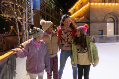 Three young female girls dressed in winter clothing on Newcastle's outdoor ice rink, with mother. All smiling and looking at one another