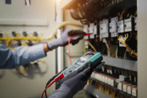 Electricity And Electrical Maintenance Service, Engineer Hand Holding AC Multimeter Checking Electric Current Voltage At Circuit Breaker Terminal And Cable Wiring Main Power Distribution Board.