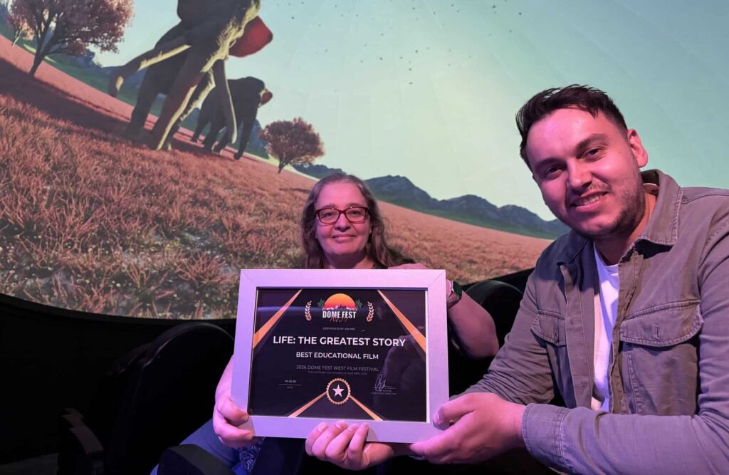 Tommy and Elin seated in Life Science Centre's Planetarium with dramatic backdrop of a herd of elephants, holding an award certificate.