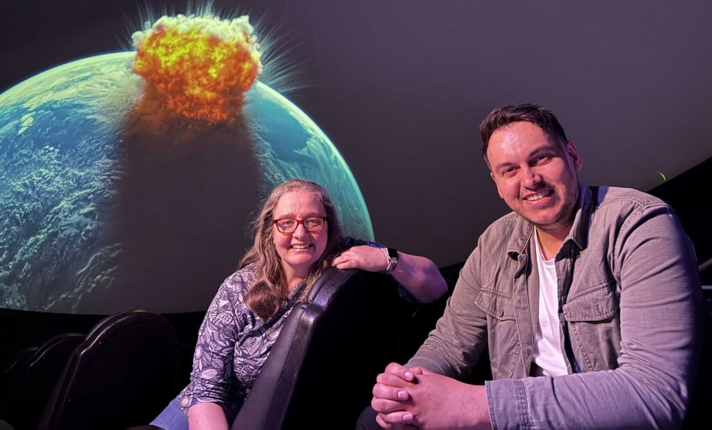 Tommy and Elin seated in Life Science Centre's Planetarium with dramatic backdrop of asteroid crashing into Earth.