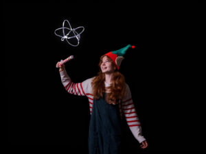 A Christmas Elf holding up a candy cane wand that makes tinsel float, she is looking up at the floating tinsel