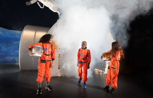 School aged students in Life Science Centre's Space Zone wearing bright orange space suits and holding helmets.
